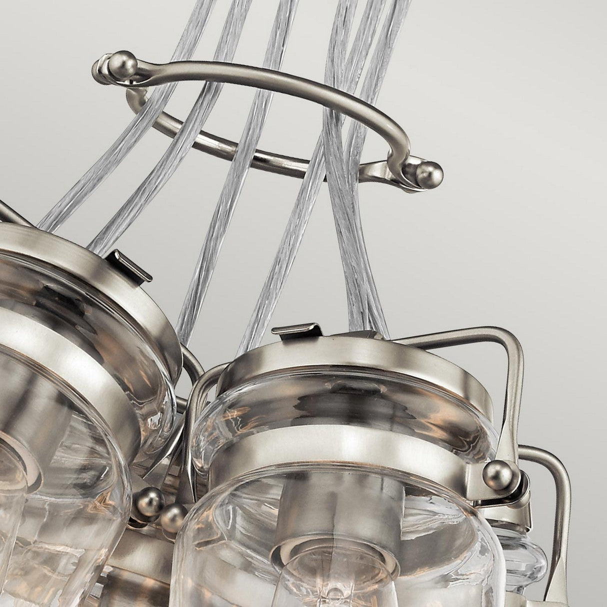 Close-up of the Brinley 6 Light Pendant in brushed nickel, showcasing clear glass shades and stylish metal fittings. The sophisticated design emanates vintage charm, with neatly gathered wires elegantly looped against a neutral backdrop.