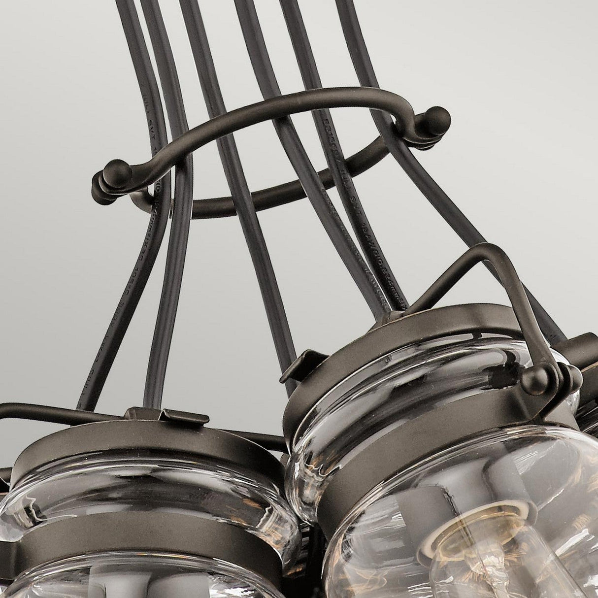 The Brinley 6 Light Pendant - Bronze is showcased in detail, presenting a rustic chandelier with a vintage-inspired design. It features several glass jar-shaped lights accented by black metal frames and curved supports, offering an elegant blend of industrial elements and a subtle bronze finish against a plain backdrop.