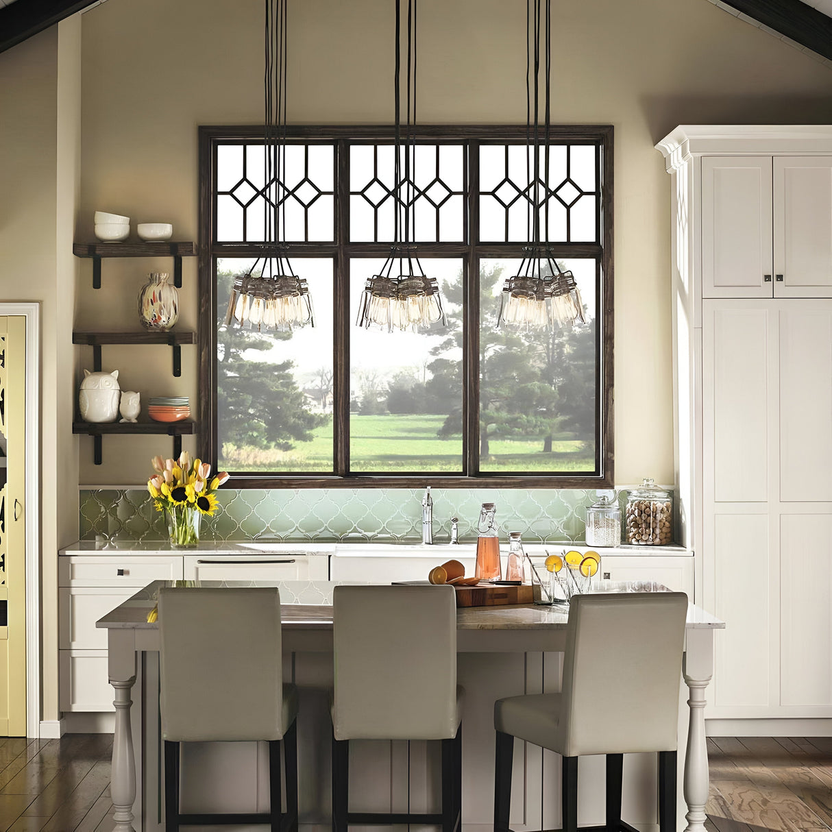 A modern kitchen with a vintage-inspired touch features a large window, the Brinley 6 Light Pendant in bronze finish, and a central island. White cabinets, shelves displaying dishes, and a glass pitcher filled with drinks complete the look, while a vase of yellow tulips adorns the countertop.