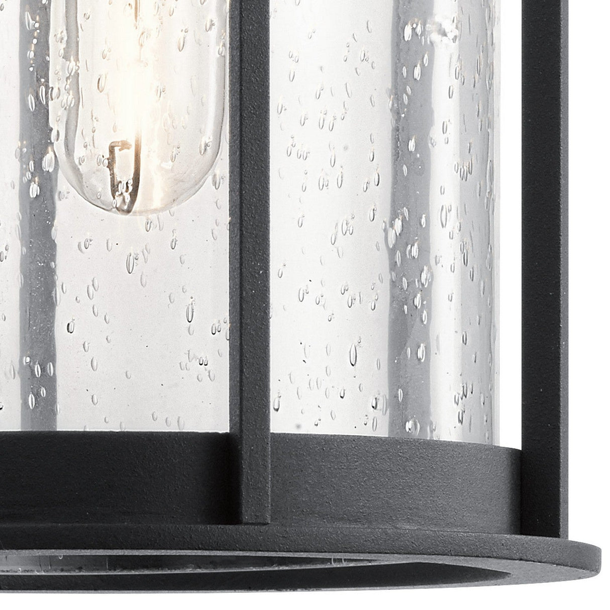 A close-up of the Camillo Outdoor 1 Light Medium Wall Lantern showcases its cylindrical clear seeded glass shade with raindrop-like textures. Inside, a slender bulb emits a soft glow. The lantern's sleek black metal frame adds a modern touch to this outdoor fixture.
