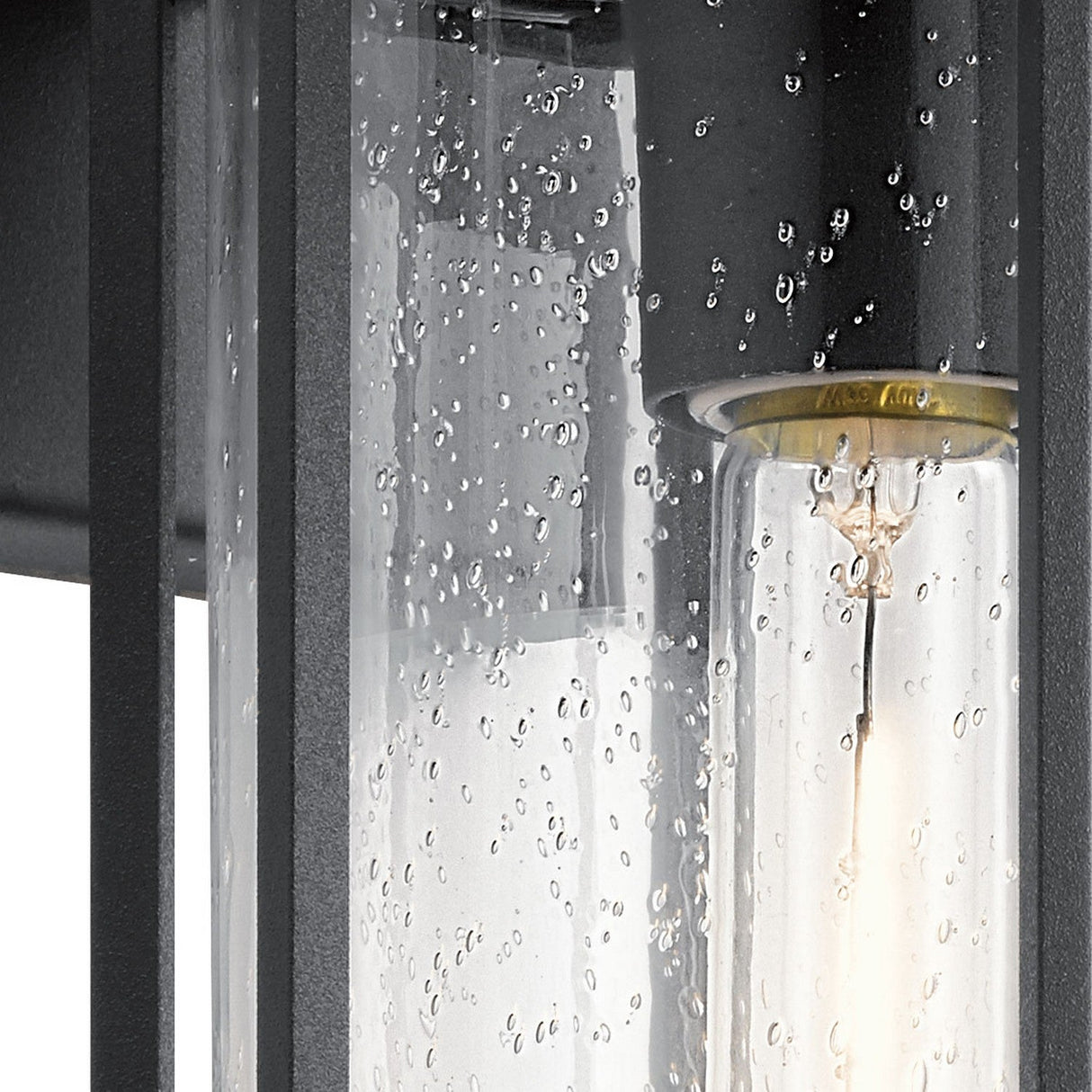 Detailed view of the Camillo Outdoor 1 Light Medium Wall Lantern showcasing its black finish and clear seeded glass panel, which creates raindrop-like patterns. The cylindrical bulb inside casts a soft glow, while the dark-colored frame adds to its modern and elegant appearance.