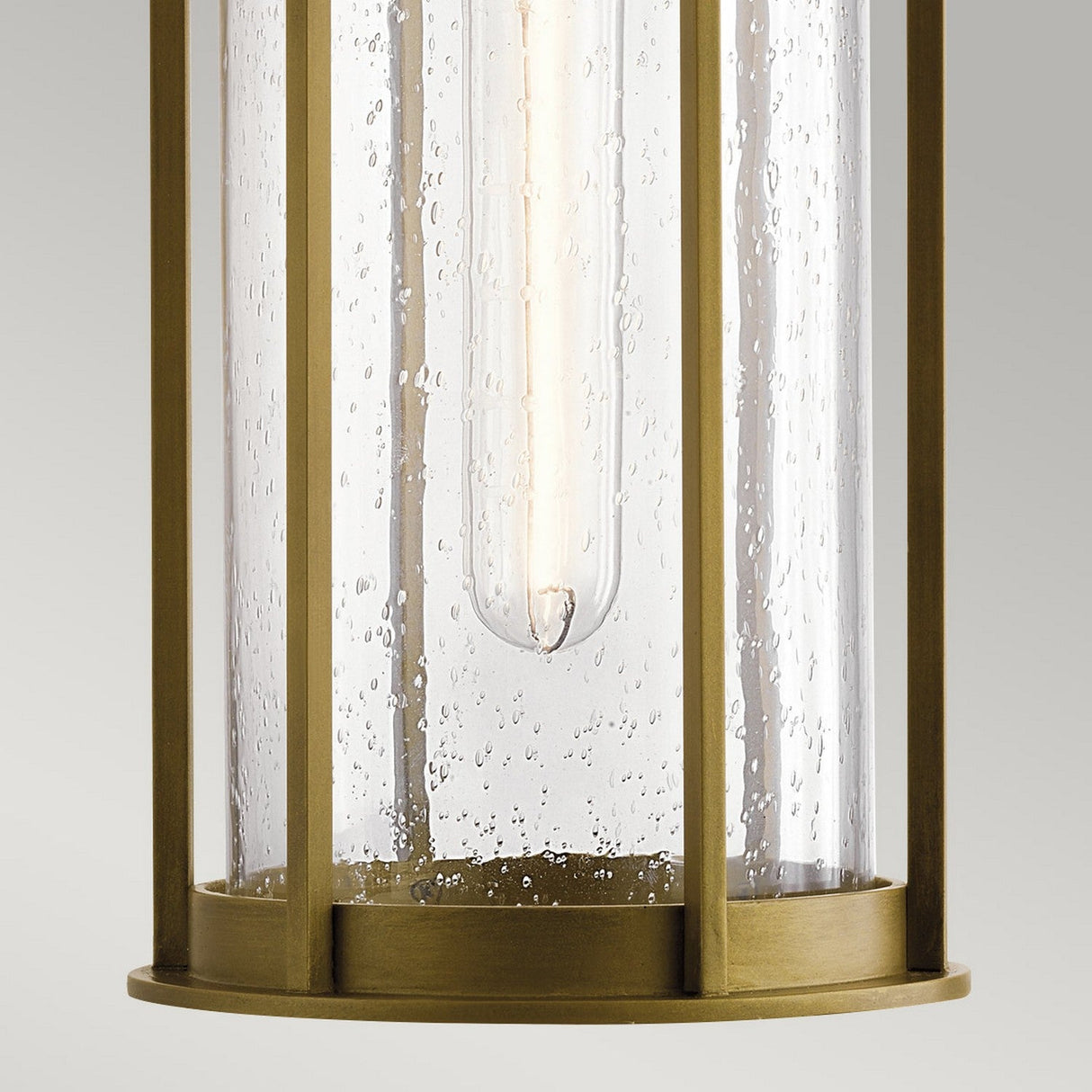 A close-up view of the Camillo Outdoor 1 Light Outdoor Pendant in natural brass reveals its cylindrical design with clear seeded glass panels and a warm LED bulb inside. The bubbled texture of the glass adds an elegant touch to this outdoor fixture.