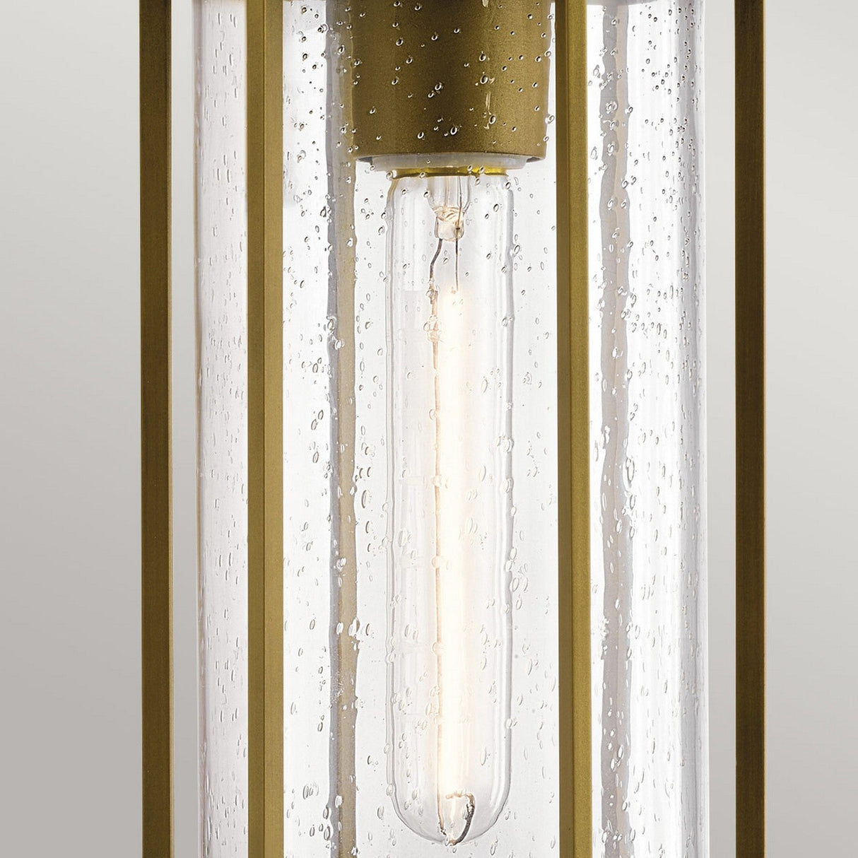 Close-up of the Camillo Outdoor 1 Light Pendant in Natural Brass, showcasing a contemporary cylindrical design. It features a long, slender bulb encased in clear seeded glass with visible bubbles, complemented by an elegant natural brass frame against a neutral background.