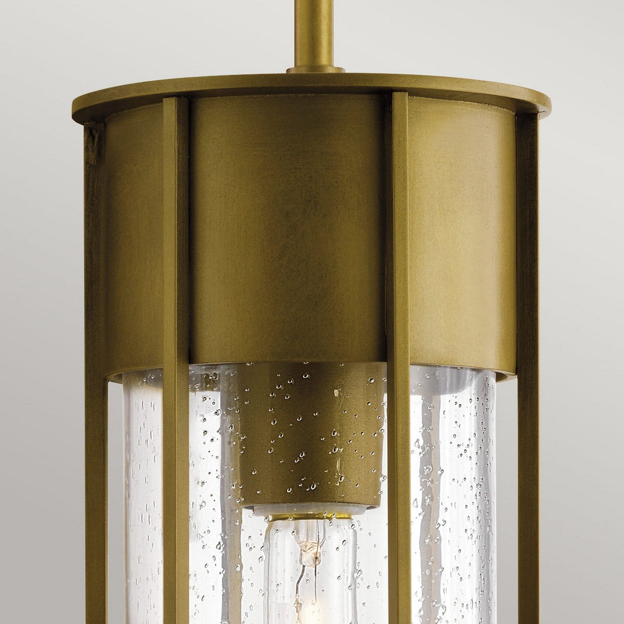 A close-up view of the Camillo Outdoor 1 Light Outdoor Pendant in natural brass, featuring cylindrical design and clear seeded glass panels with raindrop-like details. The pendant light is turned off against a plain light gray background.