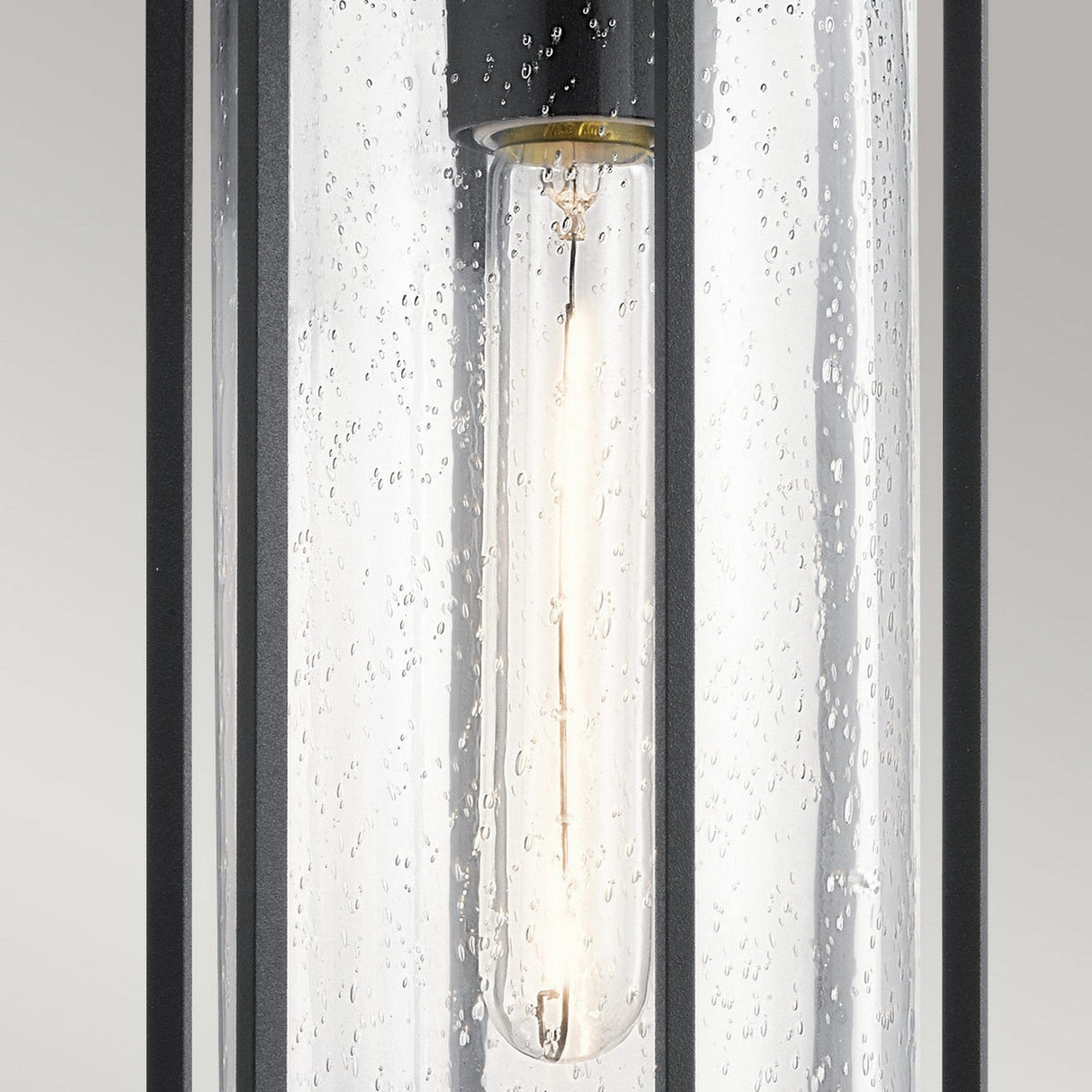 A close-up of the Camillo Outdoor 1 Light Outdoor Pendant showcases its modern cylindrical design with a clear, seeded glass cover. The elongated bulb inside emits a warm glow, while the sleek black metal frame adds a contemporary touch against a plain background.