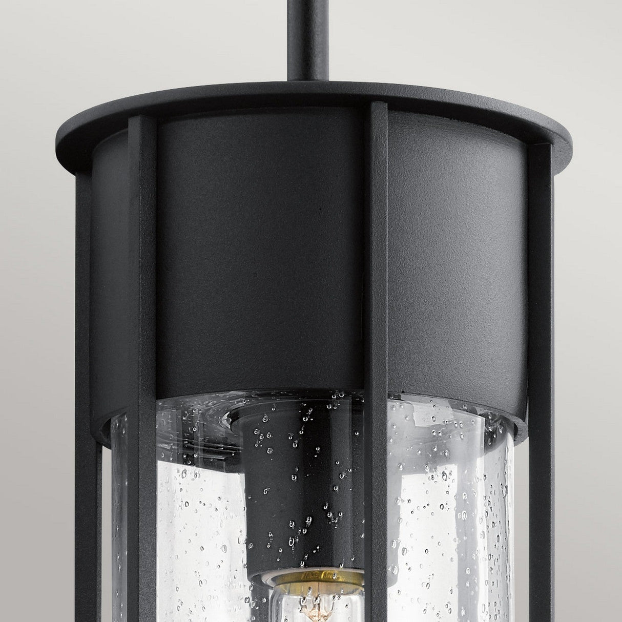 Close-up of the sleek and modern Camillo Outdoor 1 Light Pendant, featuring a cylindrical seeded glass shade encased in a black metal frame. The bubbled texture adds intrigue, revealing the bulb inside. The softly blurred background enhances its elegant design.