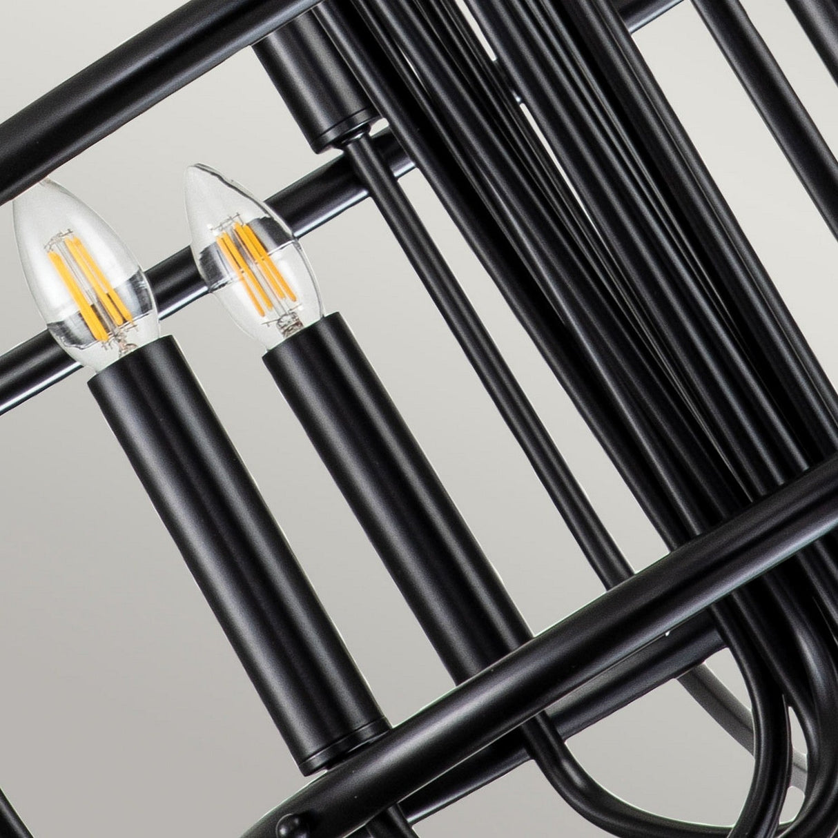 This close-up highlights the Capitol Hill 12 Light Chandelier - Black, showcasing two LED bulbs designed to resemble candles, each held in a sleek black cylindrical holder. The background reveals an arrangement of intertwined black metal rods that embody the chic and contemporary elegance of this modern lighting fixture.