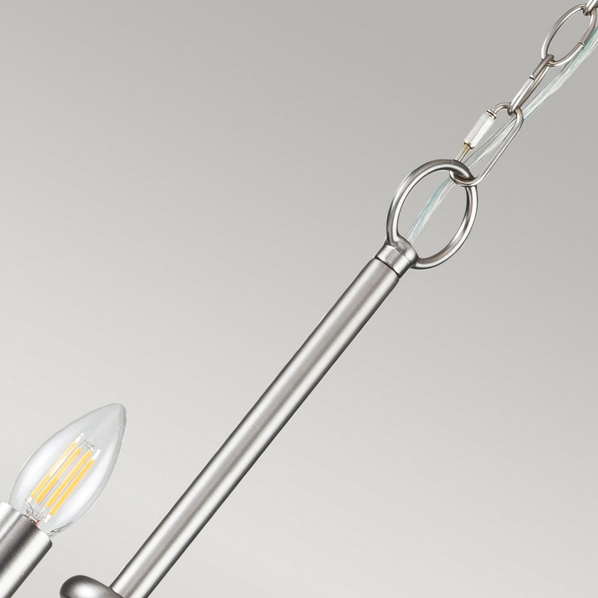 A close-up of the Capitol Hill 12 Light Chandelier in brushed nickel showcases its modern, industrial-style design. The fixture features a sleek rod and chain with candle-shaped bulbs that have clear filaments encased in metal holders, all set against a neutral background.