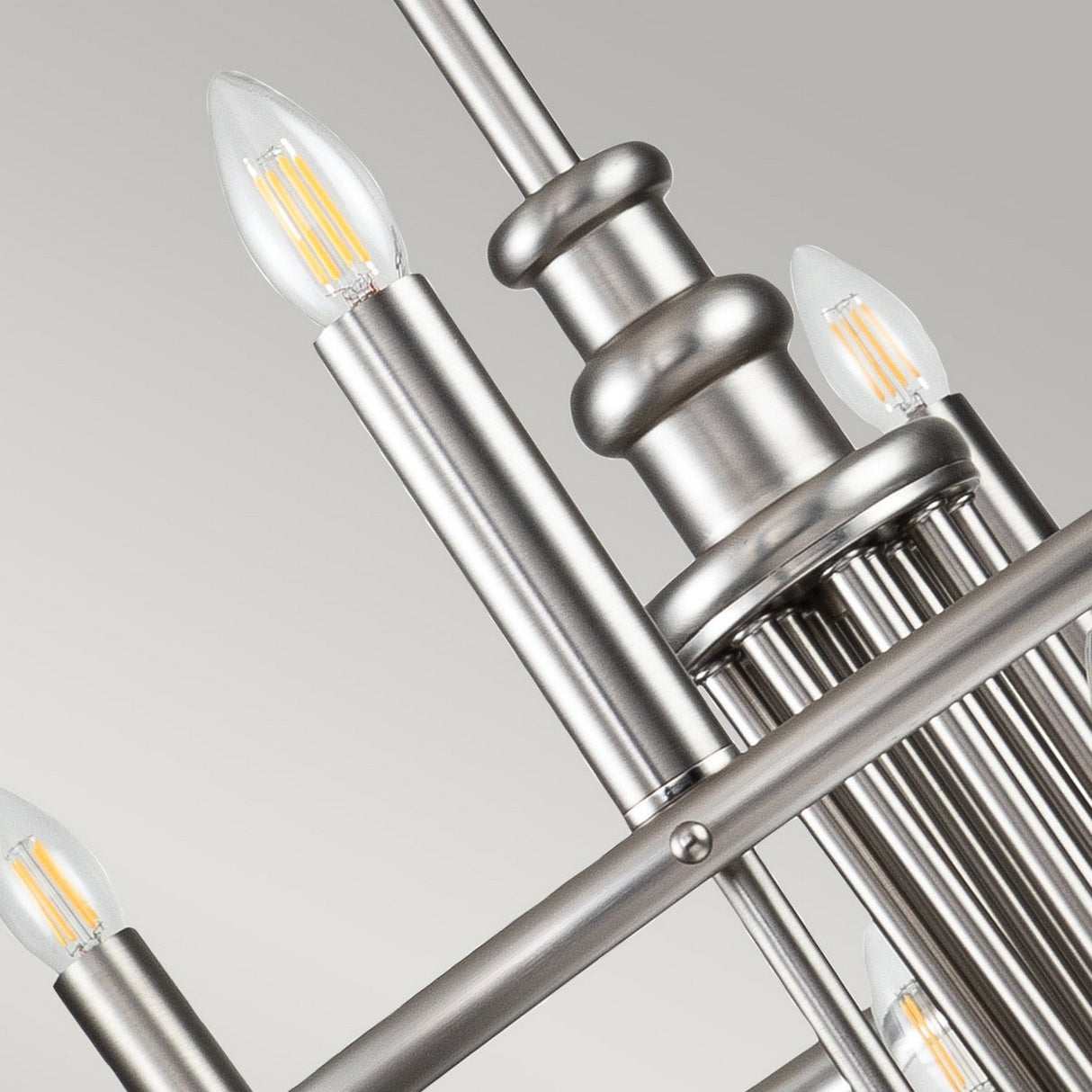 A close-up of the Capitol Hill 12 Light Chandelier in brushed nickel highlights its modern design with multiple metal rods and candle-shaped LED bulbs set against a neutral gray background.