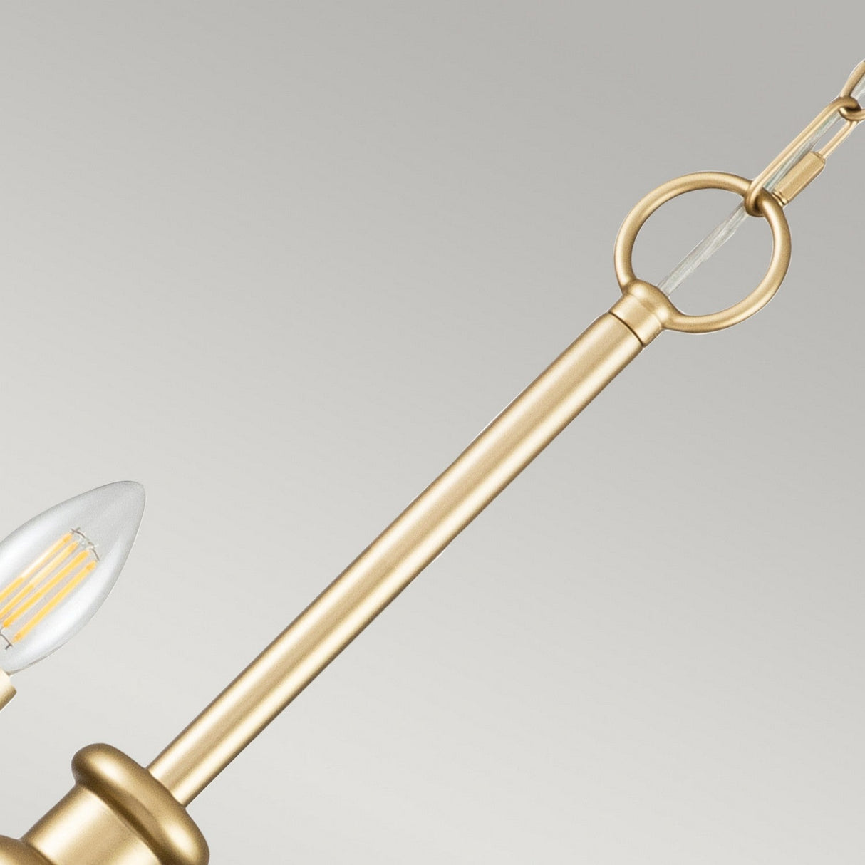A close-up of the Capitol Hill 12 Light Chandelier in brass showcases its modern design with a slender rod, circular hanging loop, and visible candle-shaped LED bulb. Its classic aesthetics enhance the plain, light background beautifully.