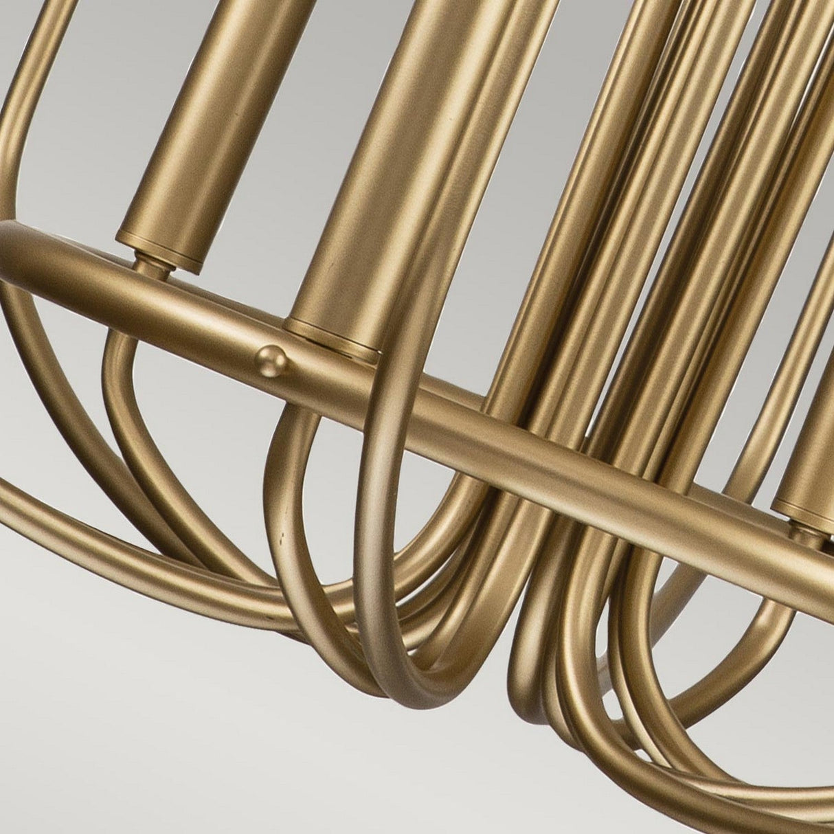 Close-up of the Capitol Hill 12 Light Chandelier - Brass, featuring a classic twelve-arm structure with gold-colored curved rods forming a geometric pattern against a grey background.