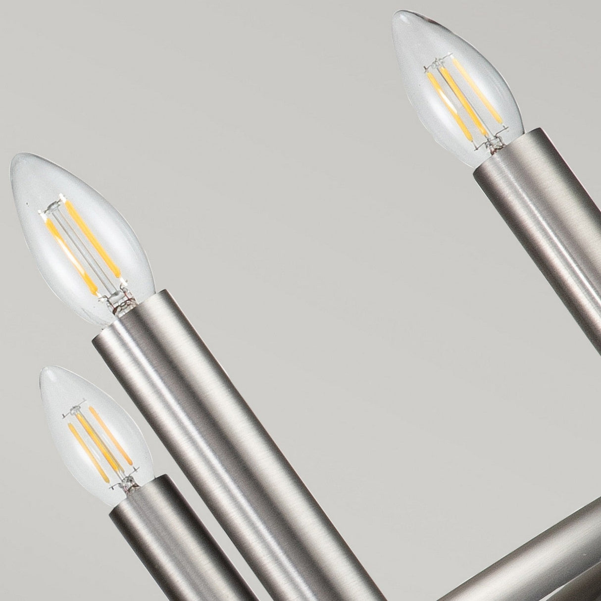 A close-up of the Capitol Hill 6 Light Chandelier in brushed nickel showcases its modern design with six candle-shaped LED bulbs elegantly placed in sleek cylindrical holders. Set against a plain background, this piece provides ambient lighting and adds a touch of contemporary flair.