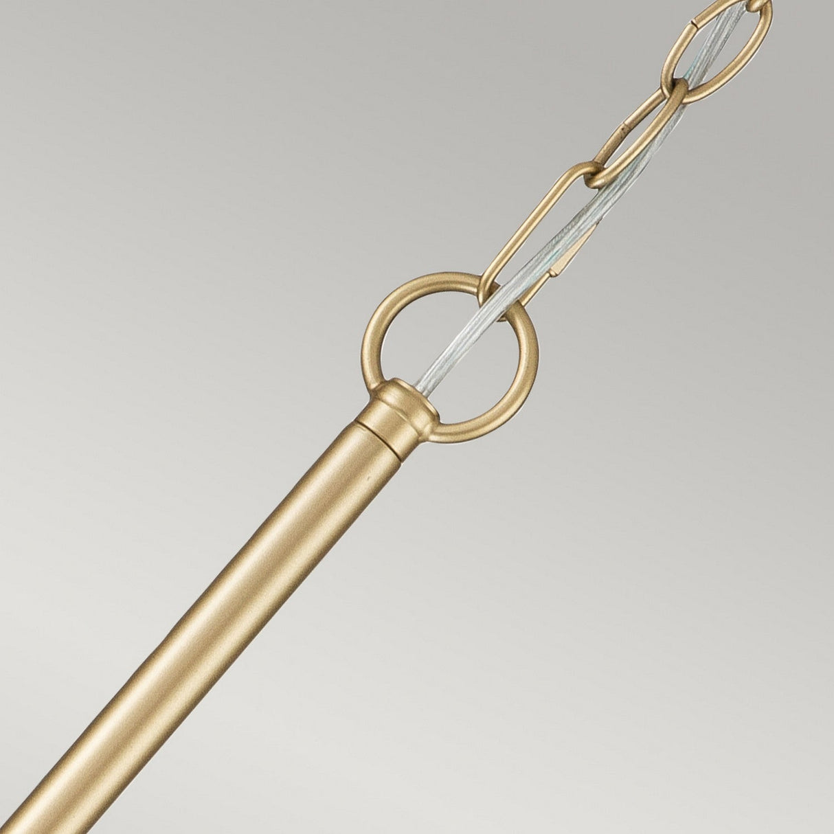 A detailed view of the Capitol Hill 6 Light Chandelier in natural brass, featuring a circular loop at the end of the rod that echoes its sophisticated design. A twisted wire cable links two metal chains, set against a simple, light gray backdrop.