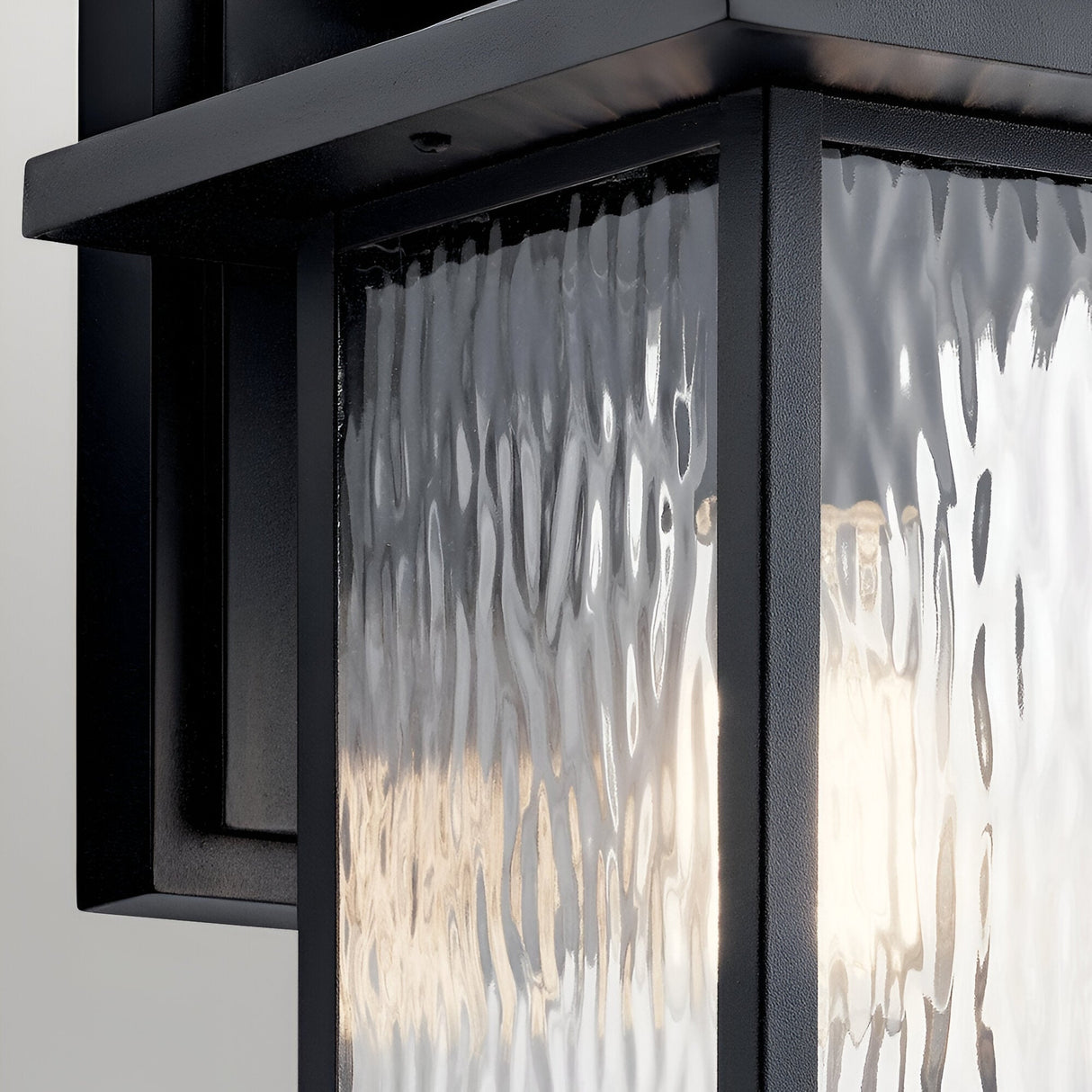 A close-up of the Cappanna 1 Light Outdoor Medium Wall Lantern in textured black, featuring a weather-resistant metal frame and waterfall glass, beautifully illuminated by a warm light inside.