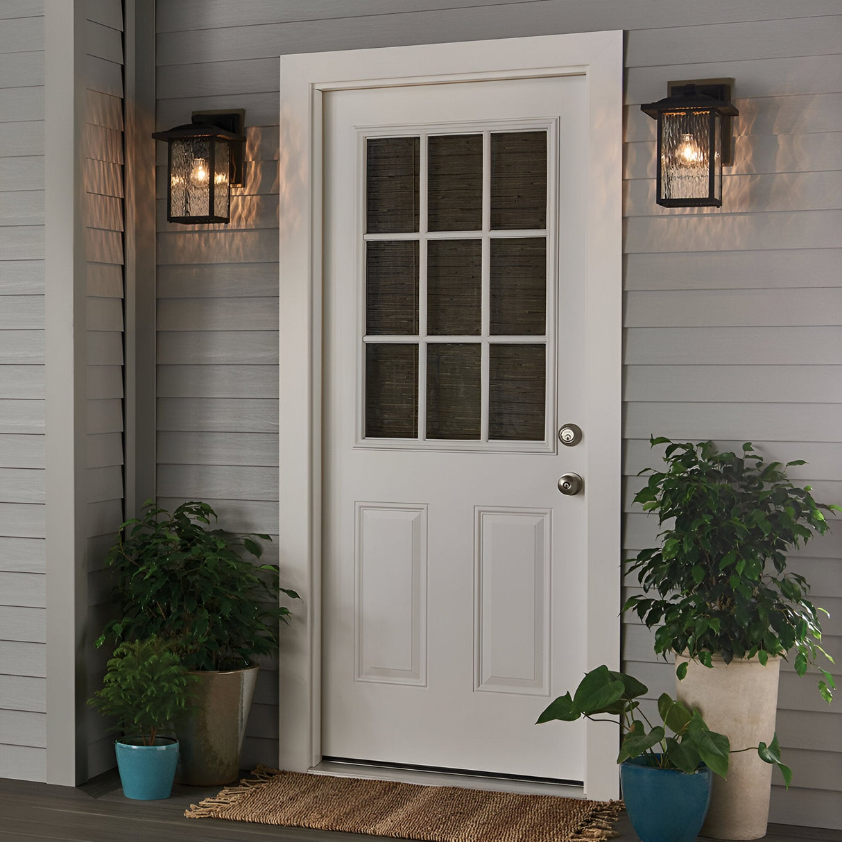 A white door with waterfall glass is centered on a gray-paneled porch, flanked by two Cappanna 1 Light Outdoor Medium Wall Lanterns in textured black. Green potted plants sit beside the door, and a brown doormat lies in front.