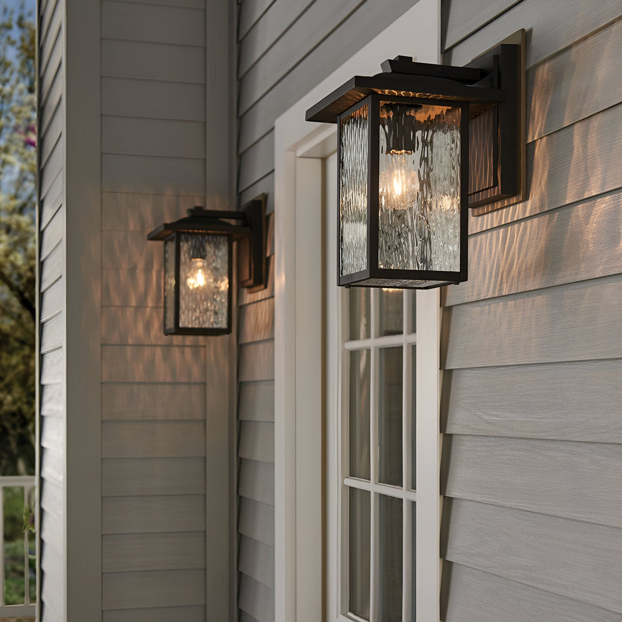 Two Cappanna 1 Light Outdoor Medium Wall Lanterns in textured black, featuring weather-resistant waterfall glass, are mounted on gray siding by a white-framed window and door. The lanterns are lit, casting patterned light.