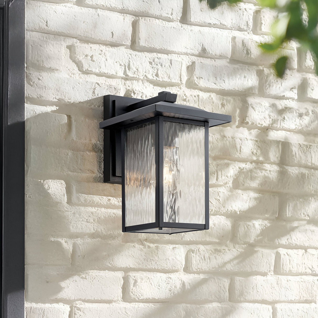 The Cappanna 1 Light Outdoor Medium Wall Lantern in textured black with weather-resistant waterfall glass is mounted on a white painted brick wall. Green leaves are partially visible in the upper right corner.
