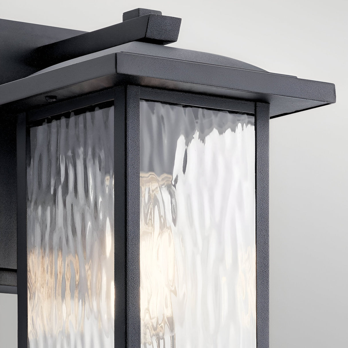 Close-up of the Cappanna 1 Light Outdoor Small Wall Lantern in textured black, featuring a sleek metal frame and waterfall glass panels, elegantly mounted on a light wall for stylish outdoor lighting.