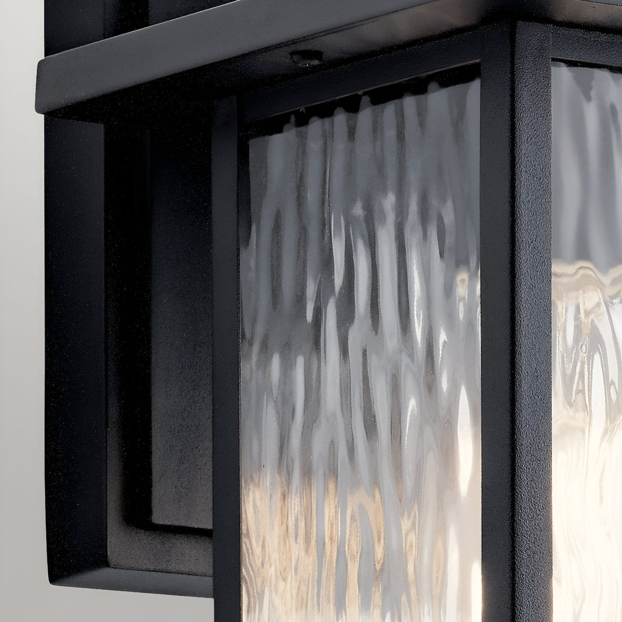 Close-up of the Cappanna 1 Light Outdoor Small Wall Lantern in textured black with a black metal frame and waterfall glass panel, showcasing elegant reflections and soft lighting—an ideal choice for outdoor spaces.