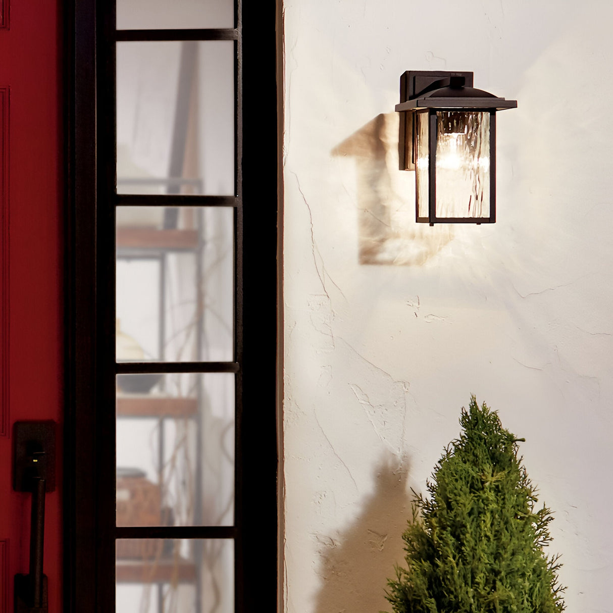 The Cappanna 1 Light Outdoor Small Wall Lantern in textured black with a waterfall glass shade provides warm, inviting light beside a red door and window, illuminating the white wall near a green shrub.