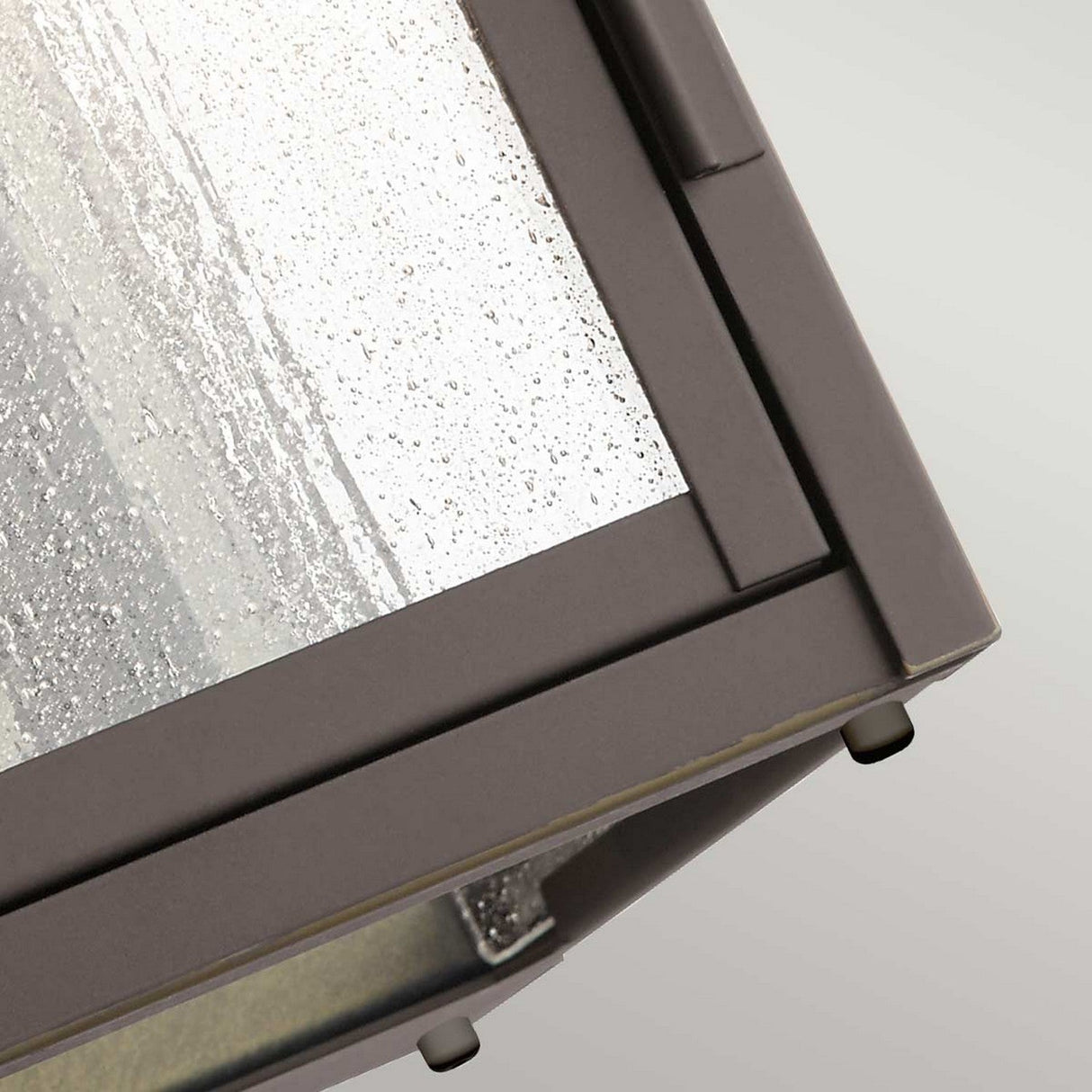 A detailed view of the Carlson Outdoor 2 Light Wall Lantern in rubbed bronze reveals a metal-framed glass corner adorned with raindrops on its seeded glass surface. The angled frame highlights the junction of two panes with condensation, evoking the style of an outdoor lantern.