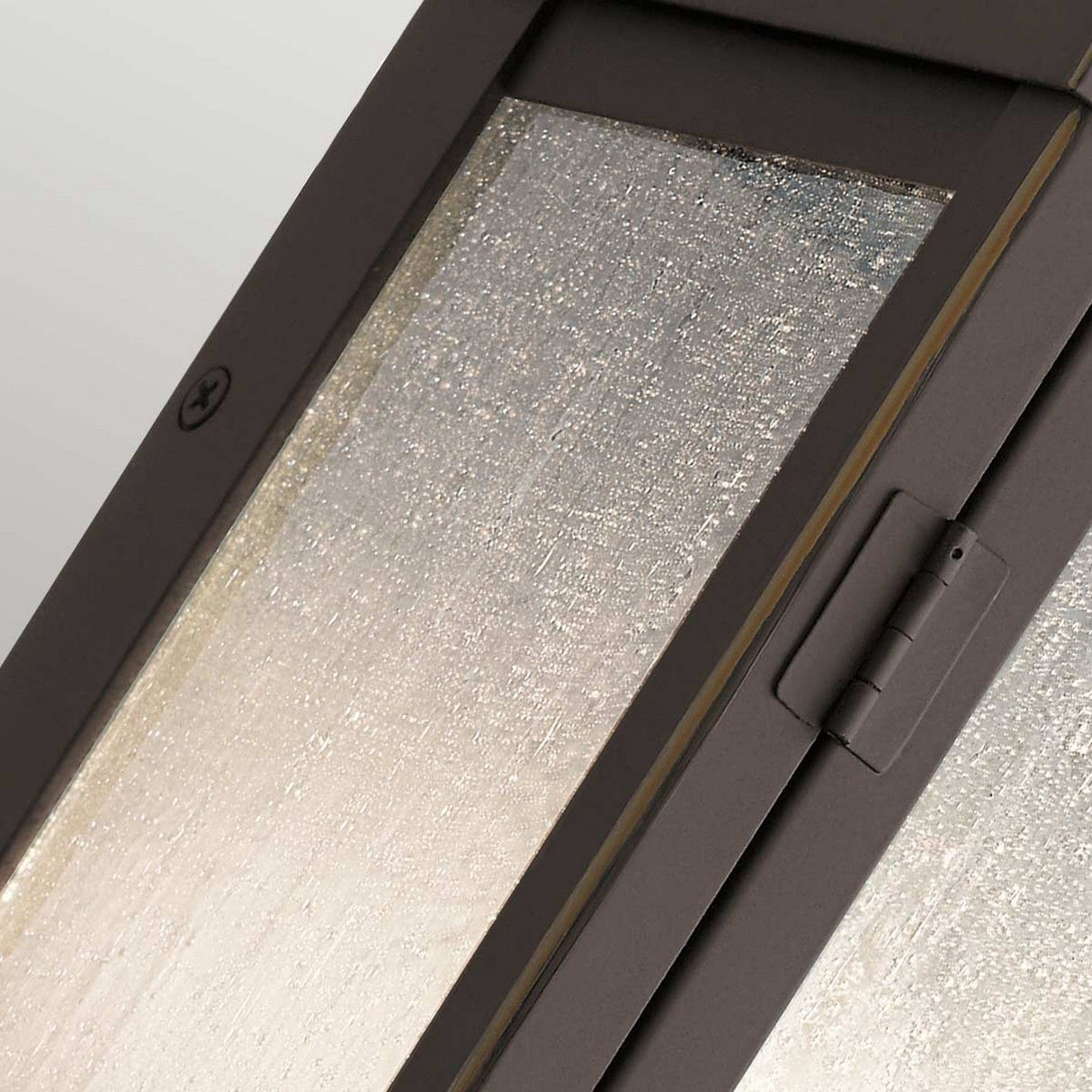 A close-up image showcases the Carlson Outdoor 2 Light Wall Lantern featuring a metal frame finished in rubbed bronze. The lantern's textured glass, reminiscent of a frosted or patterned surface similar to seeded glass, is slightly ajar, revealing the hinge and part of the frame for a modern industrial aesthetic.