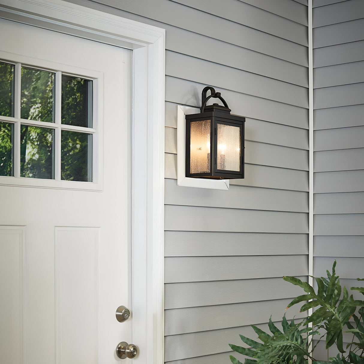 A white door with a square window sits on the left, accompanied by the Carlson Outdoor 2 Light Wall Lantern in rubbed bronze accents on a gray siding wall. A small green plant is partially visible at the bottom right.