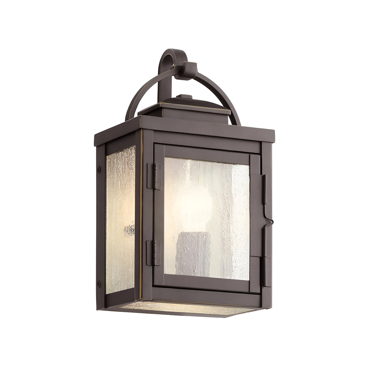The Carlson Outdoor 1 Light Wall Lantern in rubbed bronze features a rectangular design with seeded glass panes and a single light bulb. It includes a curved top handle and is stylishly mounted on a wall bracket.