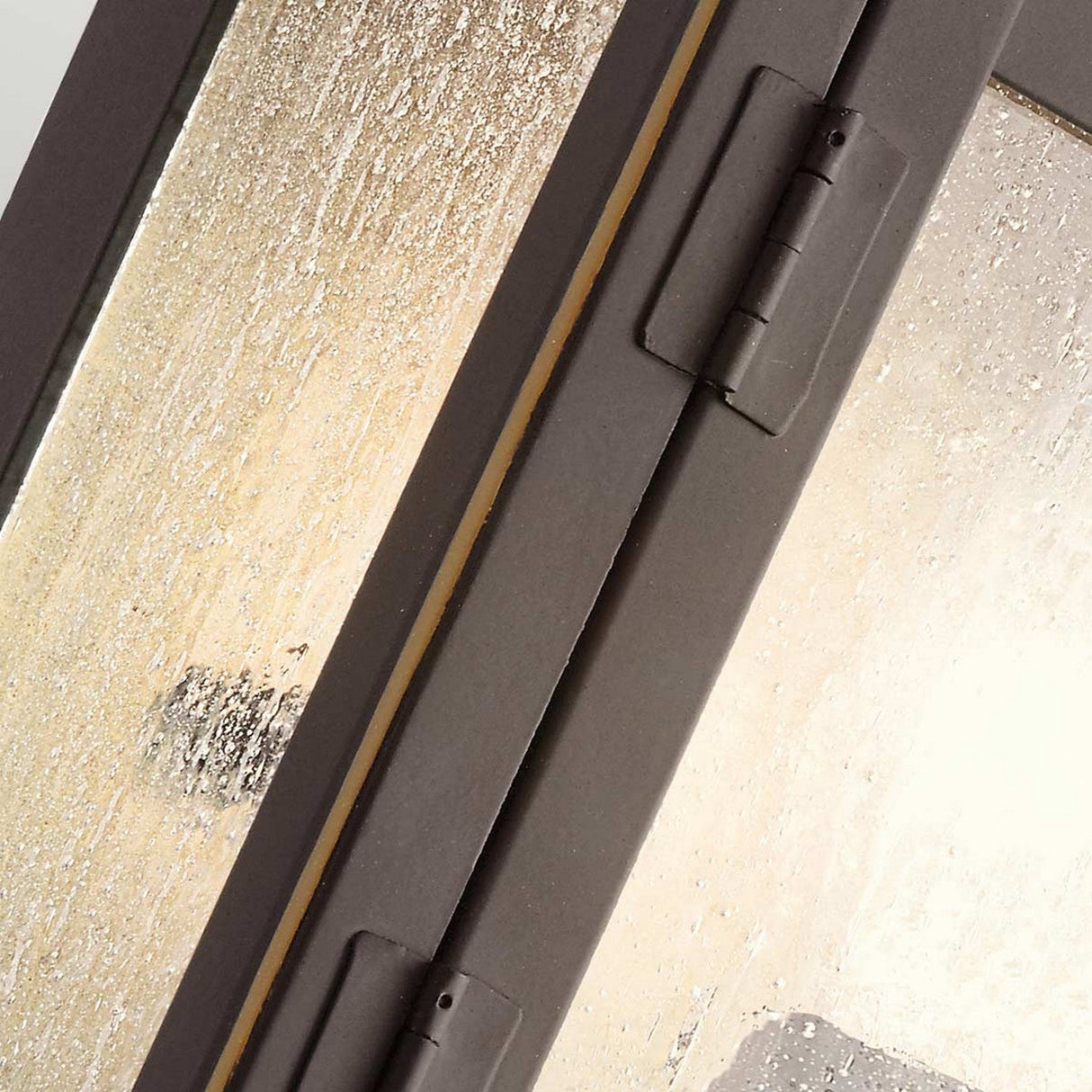 A close-up view of the Carlson Outdoor 1 Light Wall Lantern in rubbed bronze, showcasing its frosted, seeded glass. The speckled texture of the glass diffuses light elegantly through its hinged metal frame, creating a refined appearance.