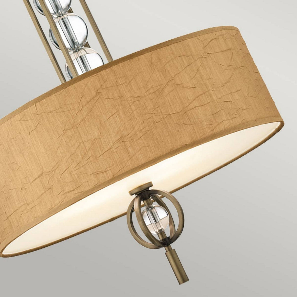 A detailed view of the Celestial 3 Light Pendant - Bronze features a beige textured drum shade adorned with optical crystal spheres inside the fixture. The design is elegantly finished with a decorative bronze metal finial, all set against a simple, light gray backdrop.