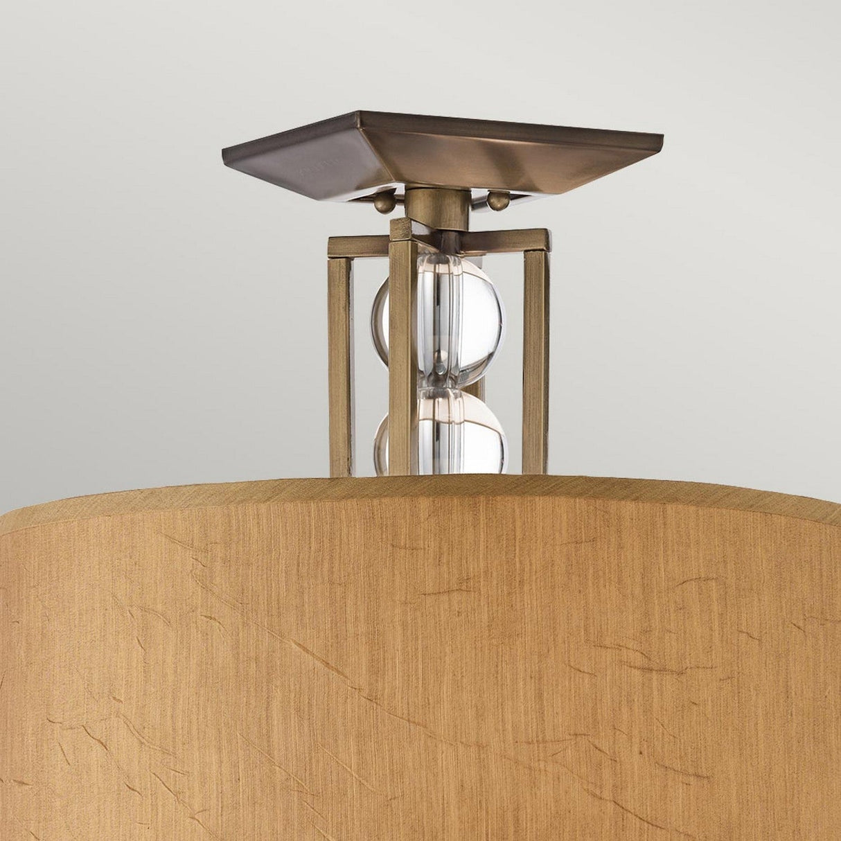 A detailed view of the Celestial 3 Light Semi Flush - Bronze reveals its textured beige lampshade. This elegant fixture boasts a bronze finish with a square metallic base, complemented by a transparent stem adorned with two decorative glass elements, all set against a plain light gray background.