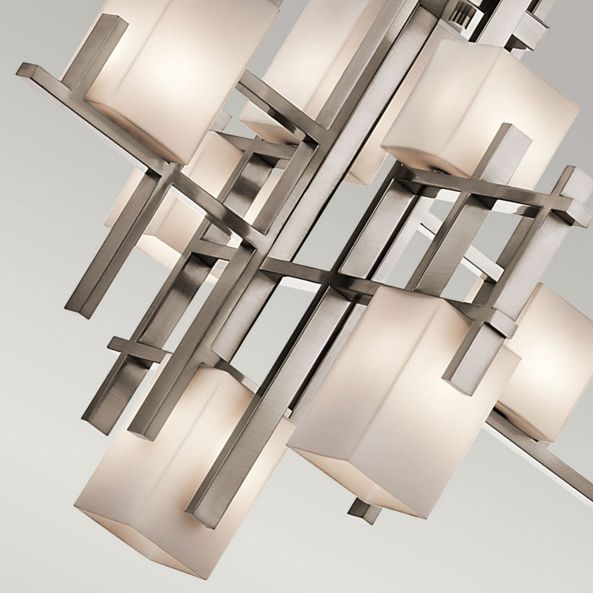 A close-up of the City Lights 7 Light Chandelier - Silver showcases its modern design with an intricate geometric pattern. The chandelier features multiple frosted square glass shades infused with LED lights, artfully arranged on metallic intersecting rods. This arrangement enhances its contemporary and artistic appearance against a neutral background.