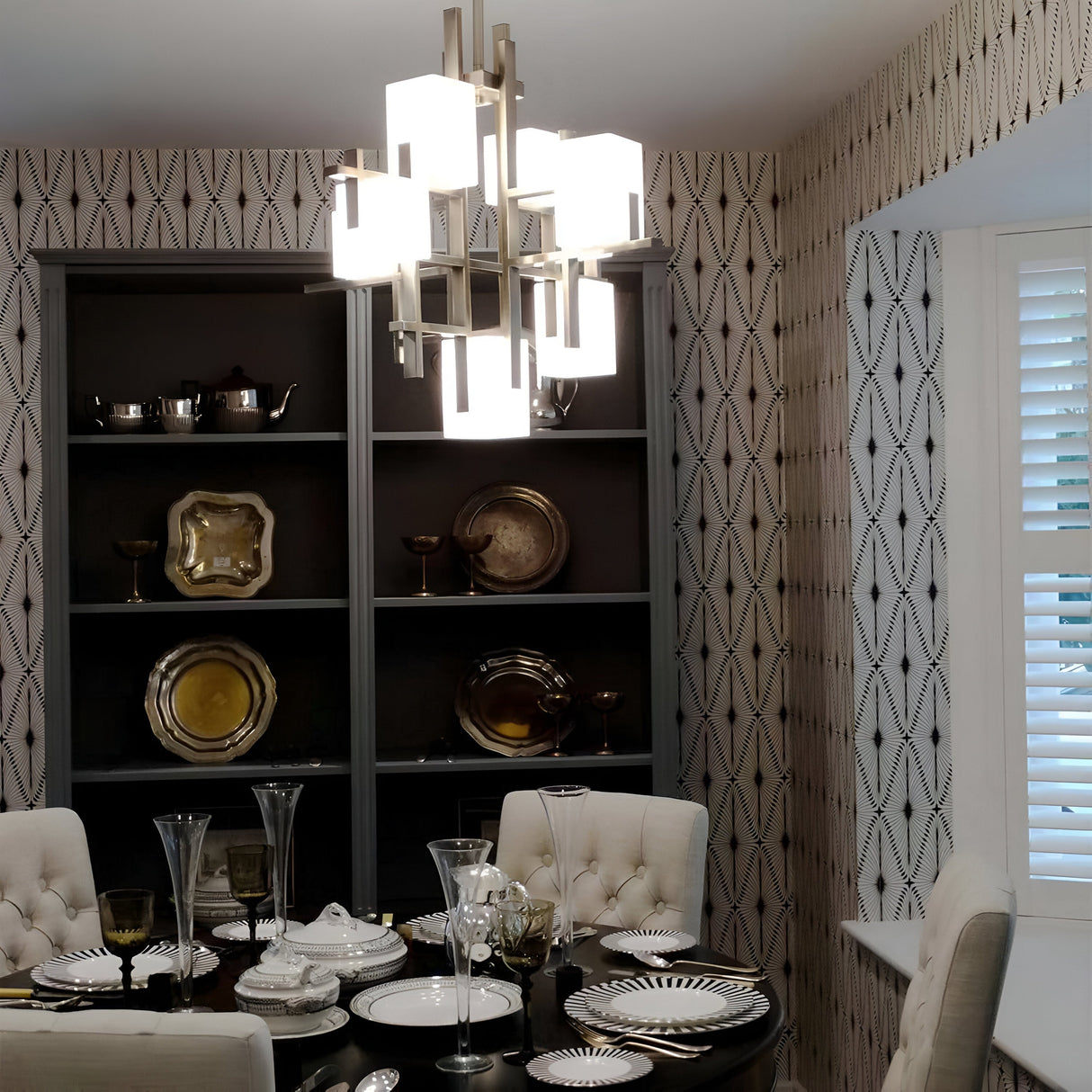 A dining room illuminated by the City Lights 7 Light Chandelier in silver, casting soft LED lights. The table is elegantly set with plates, glasses, and decorative items. The wall showcases geometric patterned wallpaper, while a shelving unit displays silver and gold dishware. Additionally, there is a window adorned with white shutters.