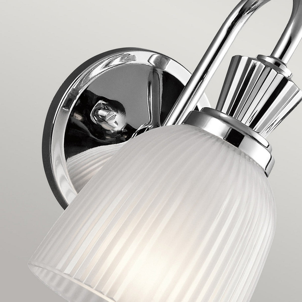 A close-up of the Cora 1 Light Wall Light showcases its ribbed, frosted glass shade and polished chrome fixture. The lamp, with an IP44 rating, is positioned against a plain light gray background.