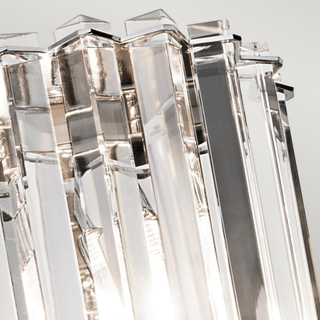 A close-up view captures a cluster of clear, vertically-oriented glass prisms with beveled edges, reflecting and refracting light to create a sparkling effect similar to that of the Crystal Skye 2 Light Wall Light - Chrome. The background is softly blurred, accentuating the sharp contours of the glass prisms.