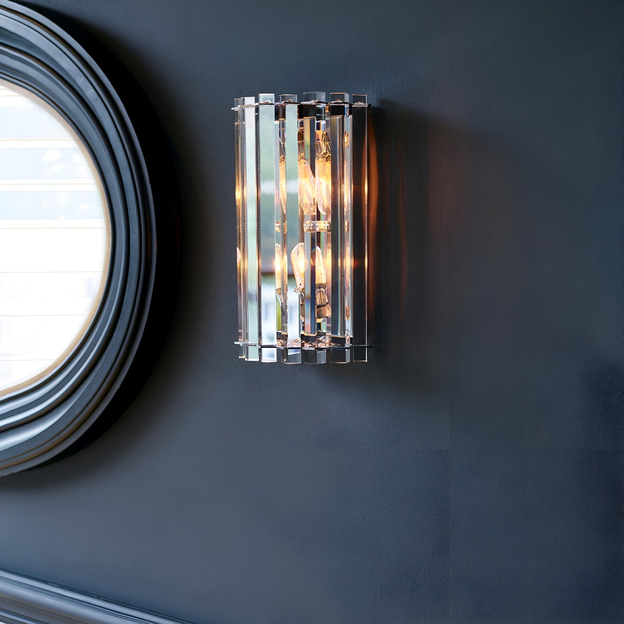 Mounted on a dark wall is the Crystal Skye 2 Light Wall Light - Chrome, featuring vertical metallic slats with a chrome finish and exposed light bulbs. In the background, a round mirror partially reflects light, enhancing the sleek and contemporary design of this piece.