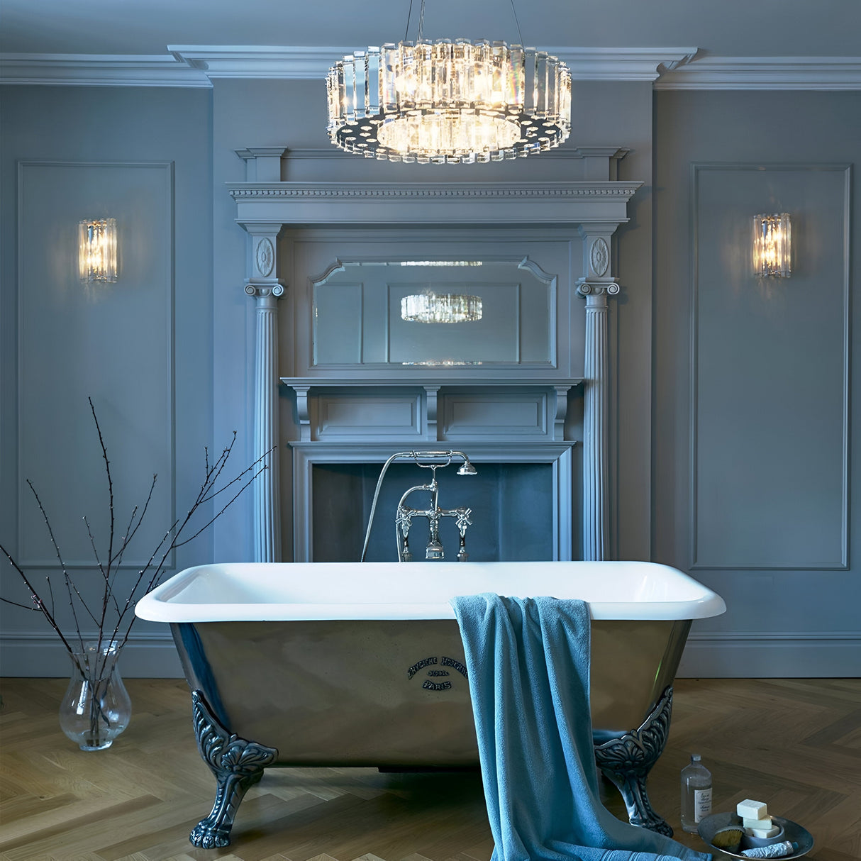 In a luxurious bathroom, a clawfoot bathtub is adorned with a blue towel draped over its side. The room, elegantly decorated in various shades of blue and featuring sophisticated wall paneling, is illuminated by the Crystal Skye 8 Light Chandelier - Chrome with its stunning triangular crystal prisms shimmering overhead. Nearby, on the wooden herringbone floor, stands a vase filled with branches.
