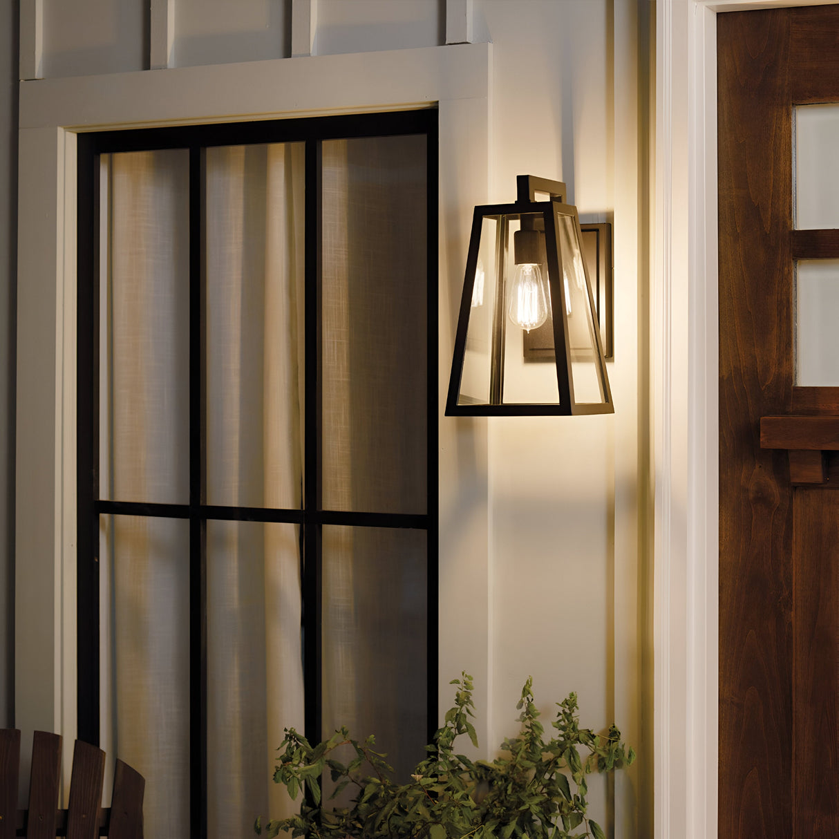 The Delison Outdoor 1 Light Wall Lantern in Rubbed Bronze (Medium), boasting an IP44 rating and a geometric design with clear glass panels and an exposed bulb, is mounted beside a window. It beautifully illuminates the wooden door and adjacent greenery, enhancing the rustic lodge exterior aesthetic.