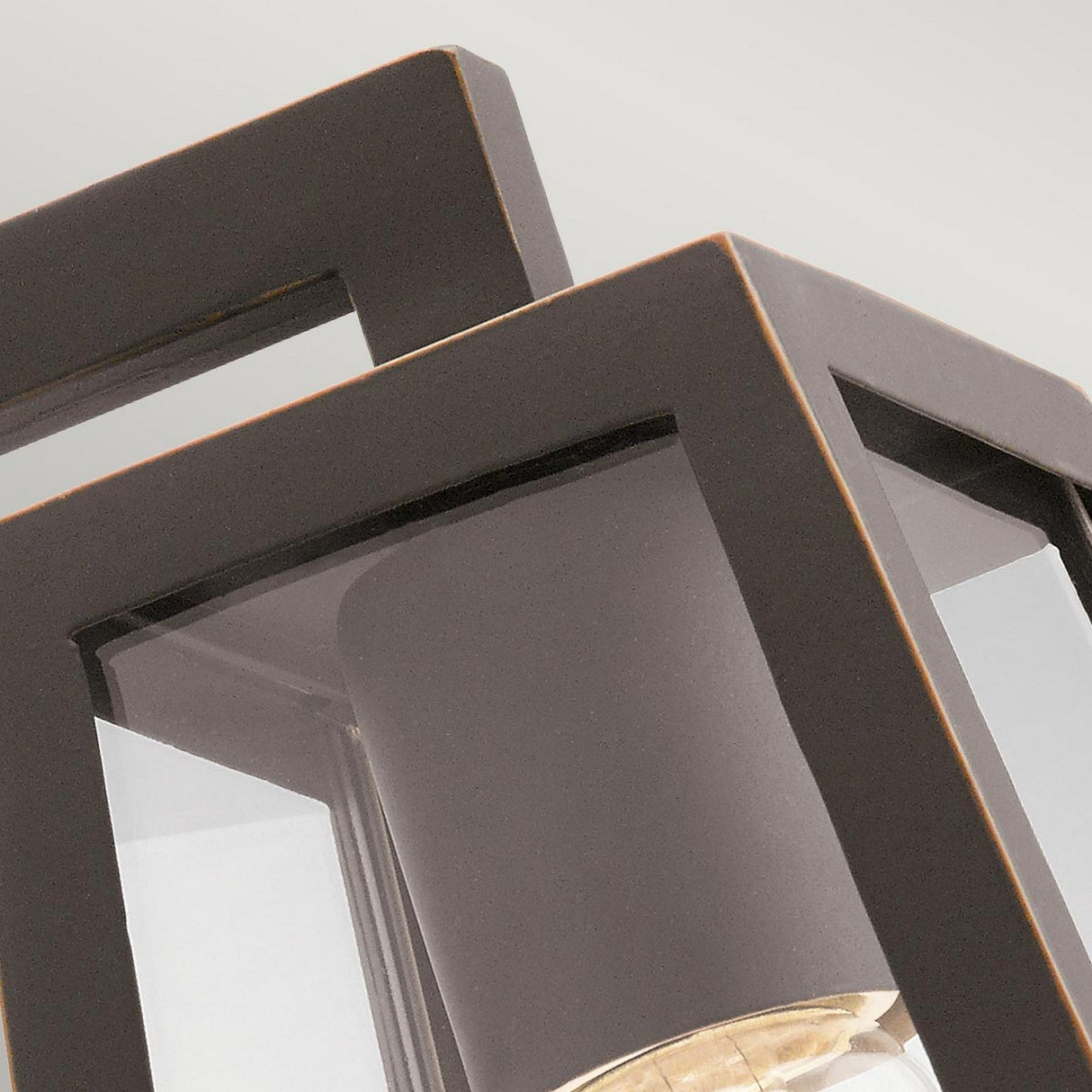 A close-up image features the Delison Outdoor 1 Light Wall Lantern in its small size, with a rubbed bronze frame and clear glass panels that reveal the bulb inside. The composition emphasizes the lantern's corners and edges against a light, neutral background.