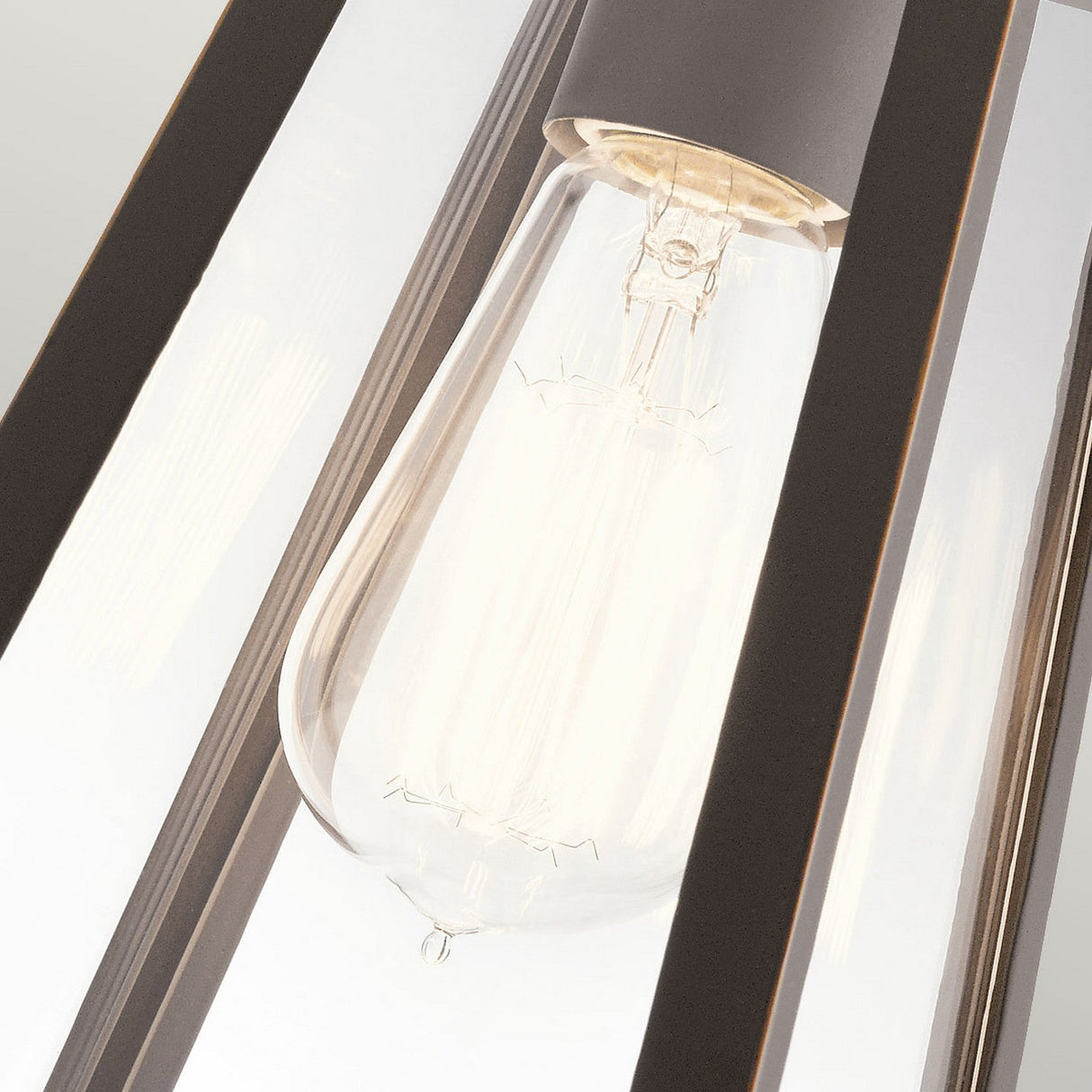 A close-up of the Delison Outdoor 1 Light Wall Lantern - Rubbed Bronze (Small), featuring a lantern-style fixture with clear glass panels and rubbed bronze metal accents. The bulb provides a warm glow that enhances its filament design, while the IP44 rating guarantees durability against outdoor elements.