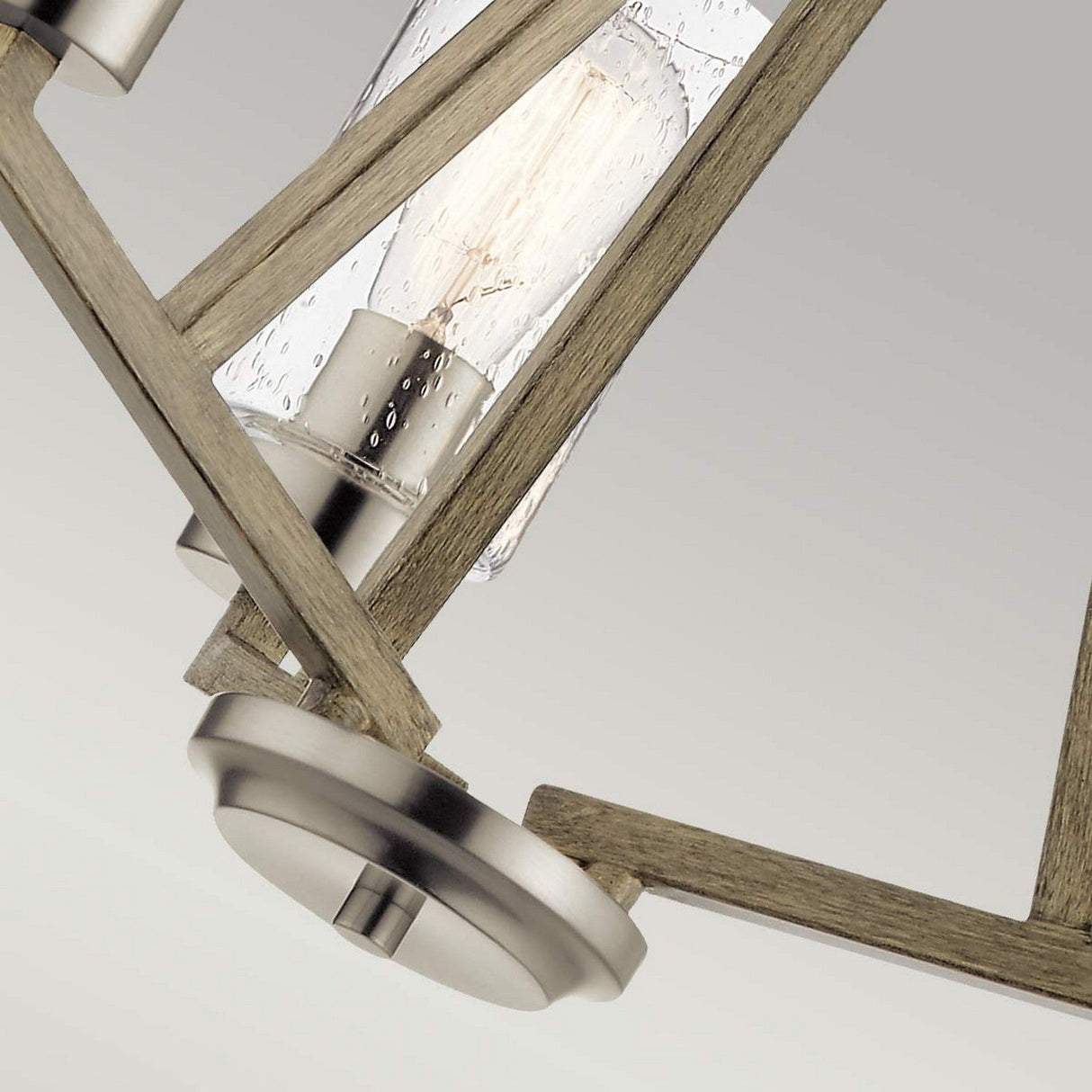 A close-up showcases the Deryn 3 Light Chandelier - Antique Grey, exuding rustic charm with its geometric design of metal and wood. The visible bulbs are elegantly encased in clear, textured glass with metal accents. An antique grey gradient in the background enhances its allure.