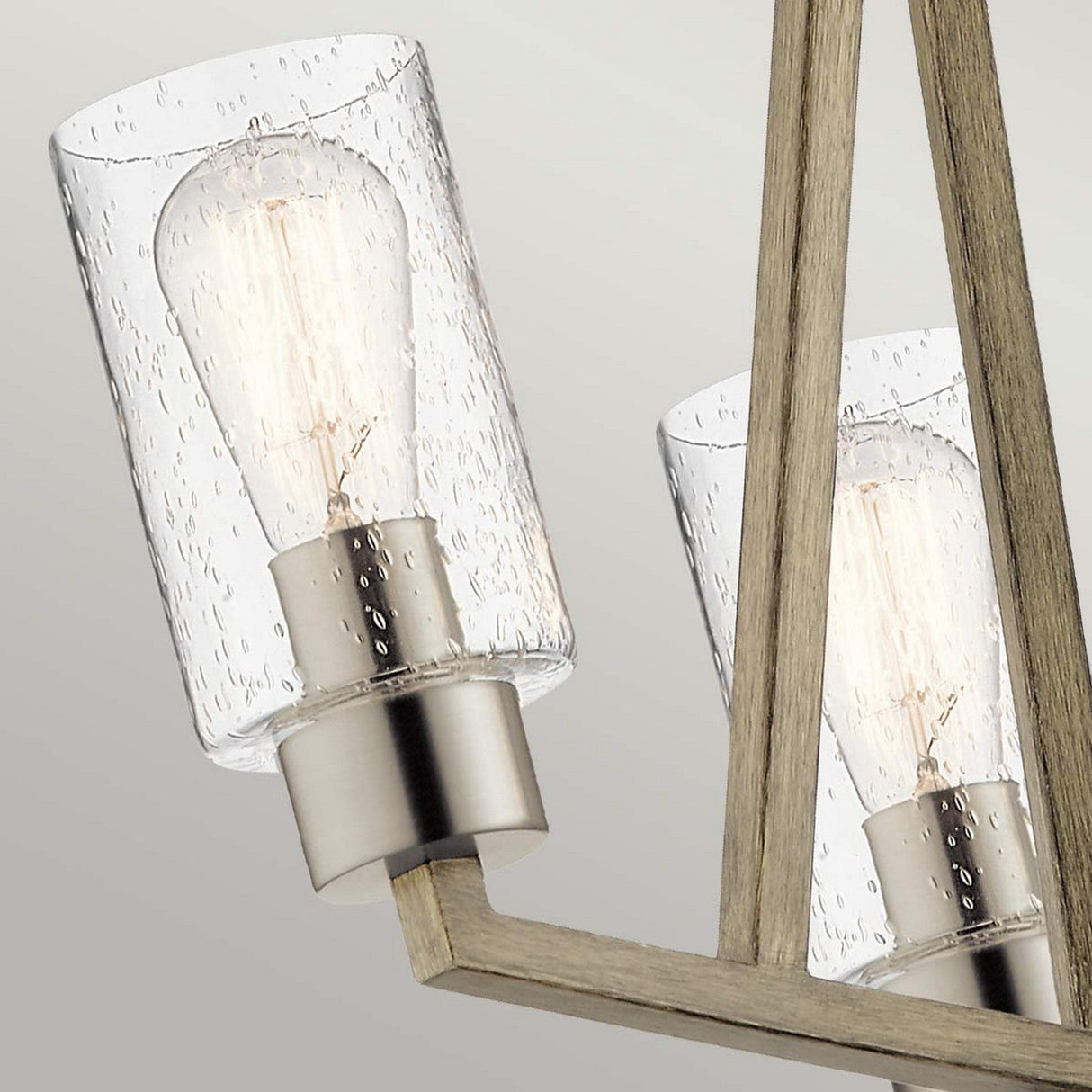 Up-close view of the Deryn 3 Light Chandelier, featuring modern light fixtures with clear seeded glass shades and exposed filament bulbs, set on brushed metal bases. The chandelier is mounted against an antique grey background.