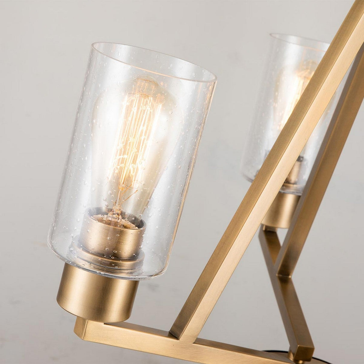 Close-up of the Deryn 3 Light Chandelier in Natural Brass, showcasing clear cylindrical glass shades and exposed filament bulbs. Supported by a gold-toned metal frame with seeded glass accents, this chandelier is set against a neutral background.