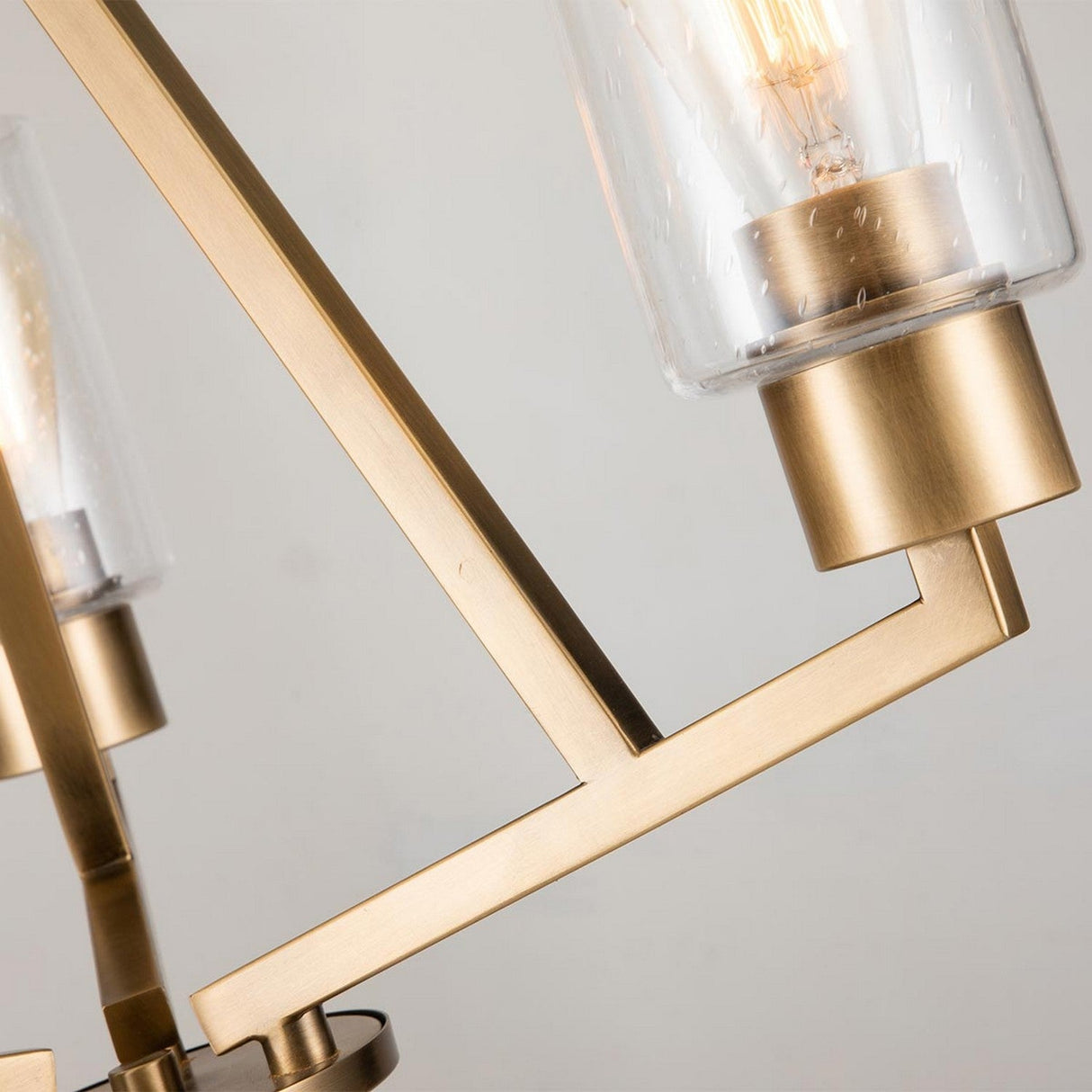 Close-up of the Deryn 3 Light Chandelier in natural brass, showcasing its angular, geometric design. This fixture boasts clear glass cylindrical shades encasing exposed Edison-style bulbs, enhancing its sleek and contemporary aesthetic.