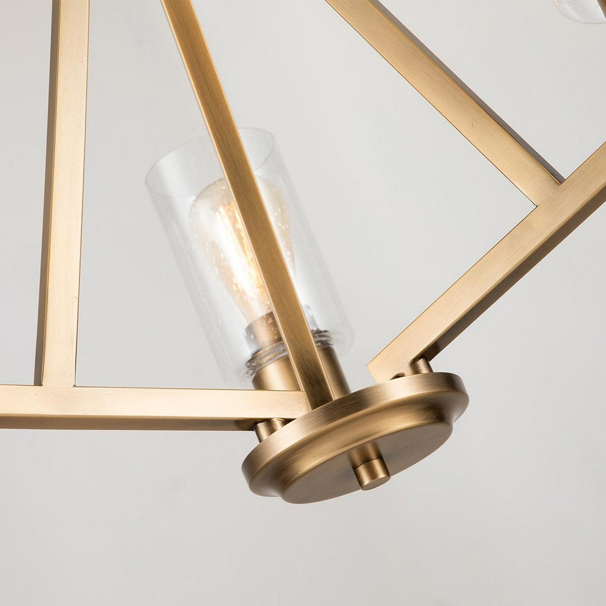 Close-up of the Deryn 3 Light Chandelier in natural brass, featuring a geometric design and a bulb cover made of seeded glass. The interior bulb emits a warm glow, enhancing its elegant style and metallic finish.