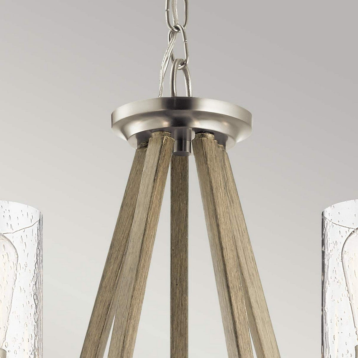 A close-up of the Deryn 5 Light Chandelier in antique grey showcases its modern design with a brushed nickel finish and rustic charm. Wooden beams converge at the top, with two speckled-textured glass shades partially visible below, complemented by accents of antique grey.