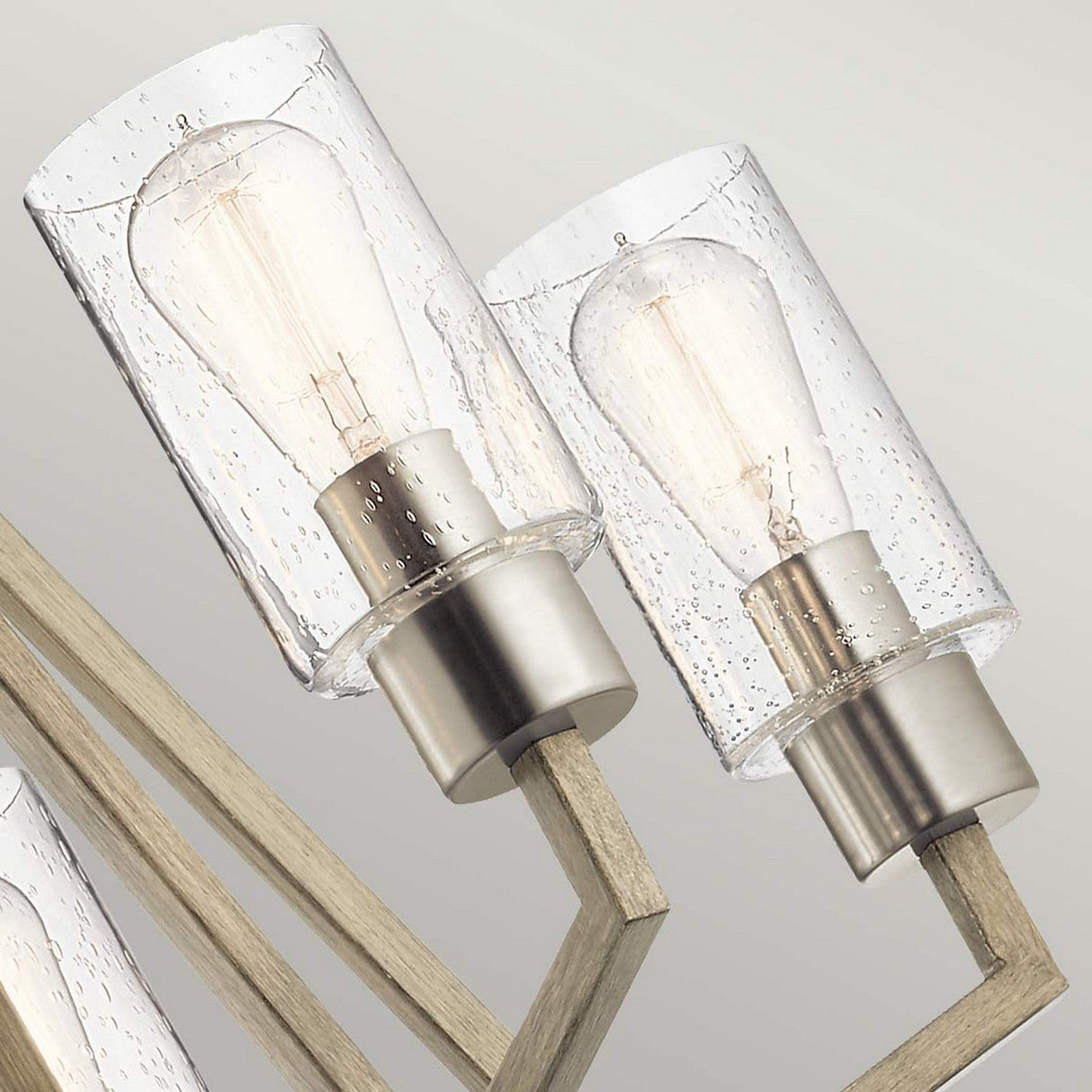 Close-up of two contemporary light fixtures featuring clear glass shades and exposed Edison bulbs. With metallic bases and wooden arms, these fixtures create a geometric pattern against a neutral backdrop, exuding a rustic charm akin to the Deryn 5 Light Chandelier in Antique Grey.