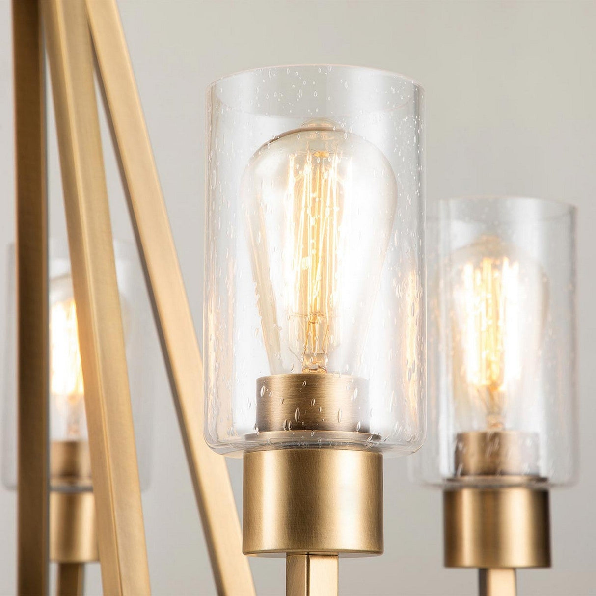 Close-up of the Deryn 5 Light Chandelier in Natural Brass featuring a clear seeded glass shade and an exposed filament bulb. The chandelier boasts a metallic, gold-toned frame that evokes rustic coastal decor, while the bulb emits a warm glow.