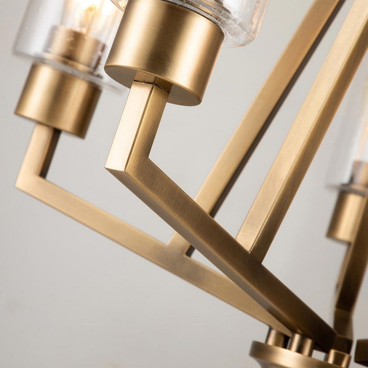 Close-up of the Deryn 5 Light Chandelier in a modern geometric design, showcasing angled metal rods with a natural brass finish and cylindrical glass shades accented with seeded glass that partially reveal light bulbs, set against a neutral background.