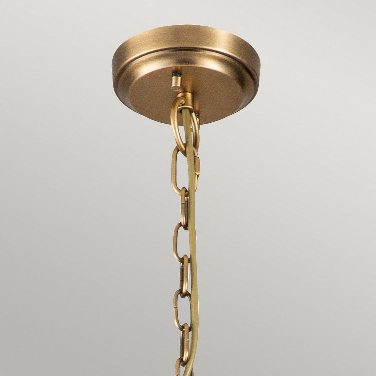 The Deryn 5 Light Chandelier in Natural Brass, featuring a brass ceiling plate with an attached chain, hangs against a light gray background, evoking the charm of rustic coastal decor. The chain is composed of interlinked oval loops that enhance its elegance with a metallic sheen, reminiscent of the chandelier's natural brass finish.
